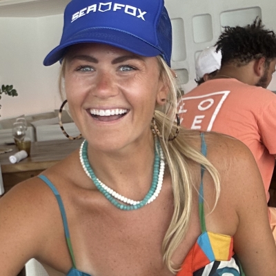 Harlan Renken headshot. A tan white woman with bland hair pulled back in a ponytail wearing a blue baseball hat. She wears a dark green patterned spaghetti strap top. She smiles very happily. 